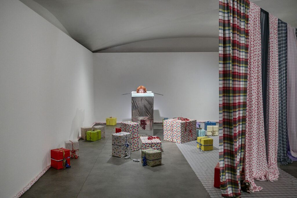Scena della performance: pacchi regalo colorati a terra, una figura su un piedistallo bianco e tende a quadri rosse e blu