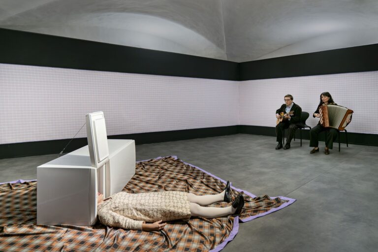 Performance alla Strozzina: una performer sdraiata su una coperta a quadri, dietro due musicisti con chitarra e fisarmonica