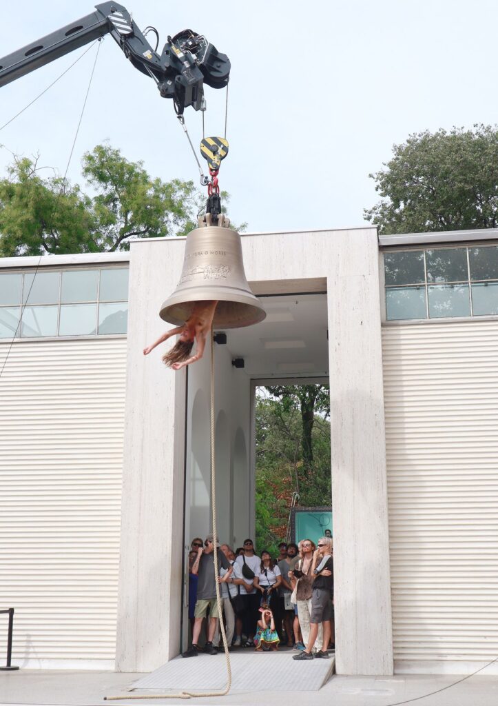Una grande campana sollevata da una gru davanti a un padiglione bianco, con una performer appesa e il pubblico sotto