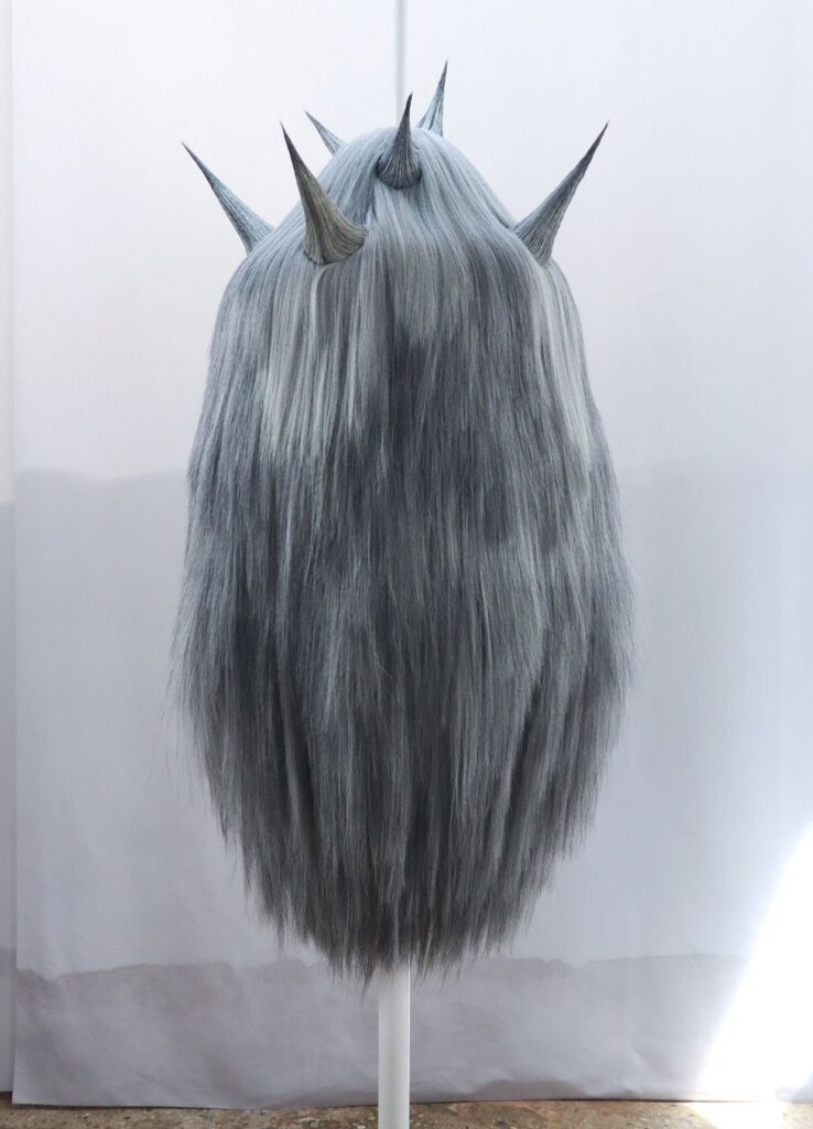 Una figura ovale coperta di lunghi capelli grigi e coronata di spine, montata su un’asta davanti a un fondo bianco