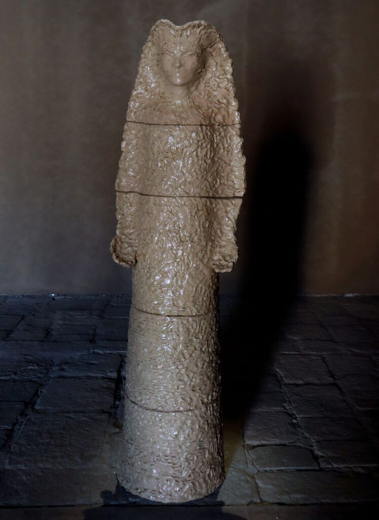 Una figura femminile alta color sabbia, tutta pieghe e drappeggi, in piedi in una sala buia dal pavimento di pietra