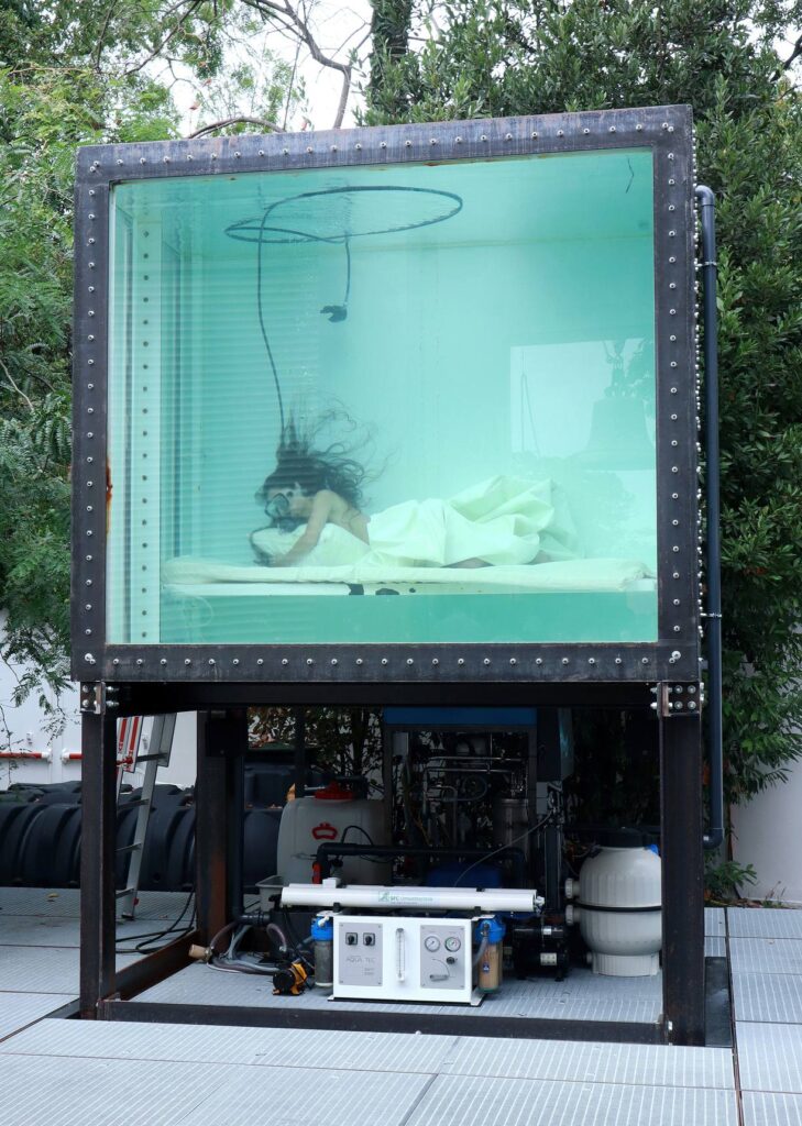 Una performer immersa in una grande vasca d’acqua trasparente, distesa su un letto sotto un lenzuolo, fra gli alberi