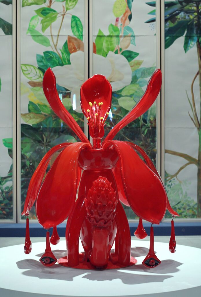 Una scultura rossa lucida con forme di fiore e di animale, su un piedistallo bianco davanti a un paravento dipinto