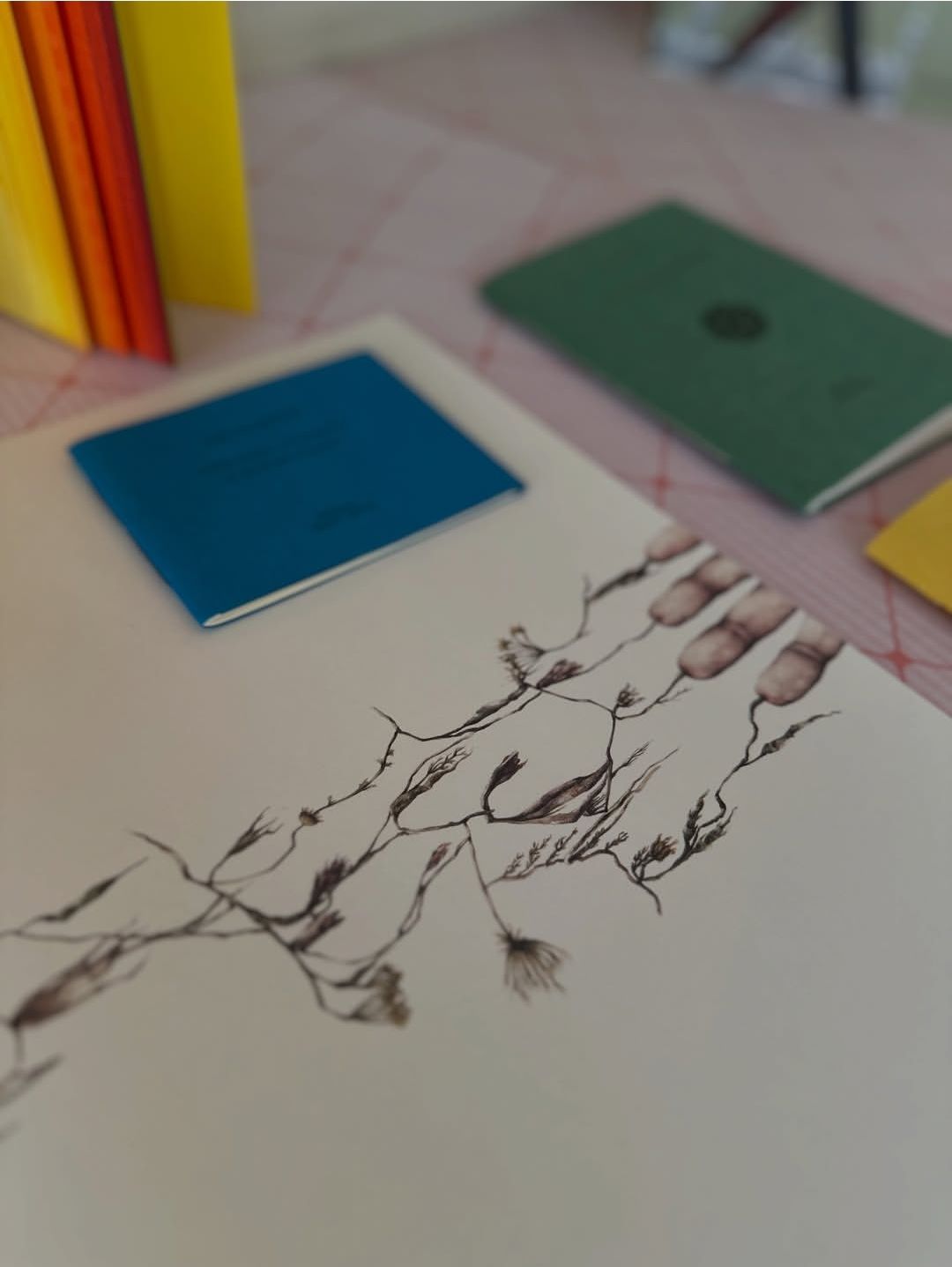 Foglio bianco con un disegno a china di rami, accanto a piccoli quaderni blu e verdi su un piano rosa