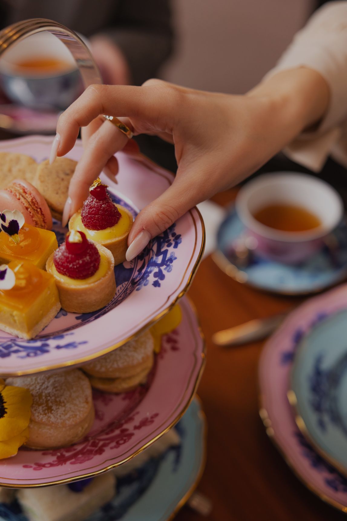 A hand picks up a mixed-berry tartlet from a Ginori porcelain cake stand, next to a cup of tea