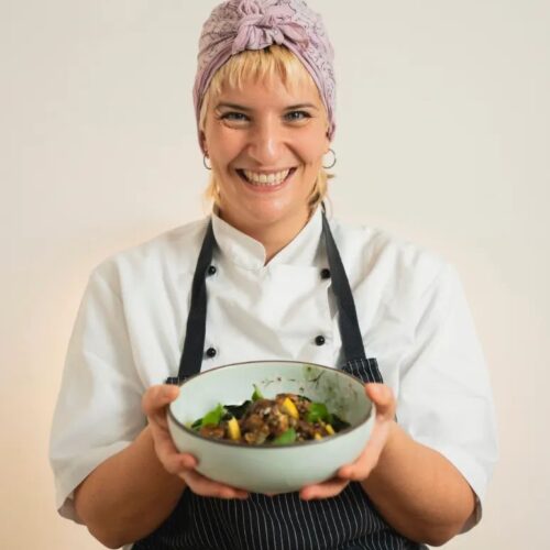 Una cuoca sorridente con turbante lilla, giacca bianca e grembiule scuro porge una ciotola di verdure e legumi