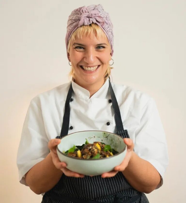 Una cuoca sorridente con turbante lilla, giacca bianca e grembiule scuro porge una ciotola di verdure e legumi