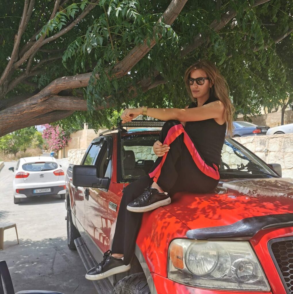 Una donna con occhiali da sole seduta sul cofano di un pick-up rosso, all’ombra di un grande albero