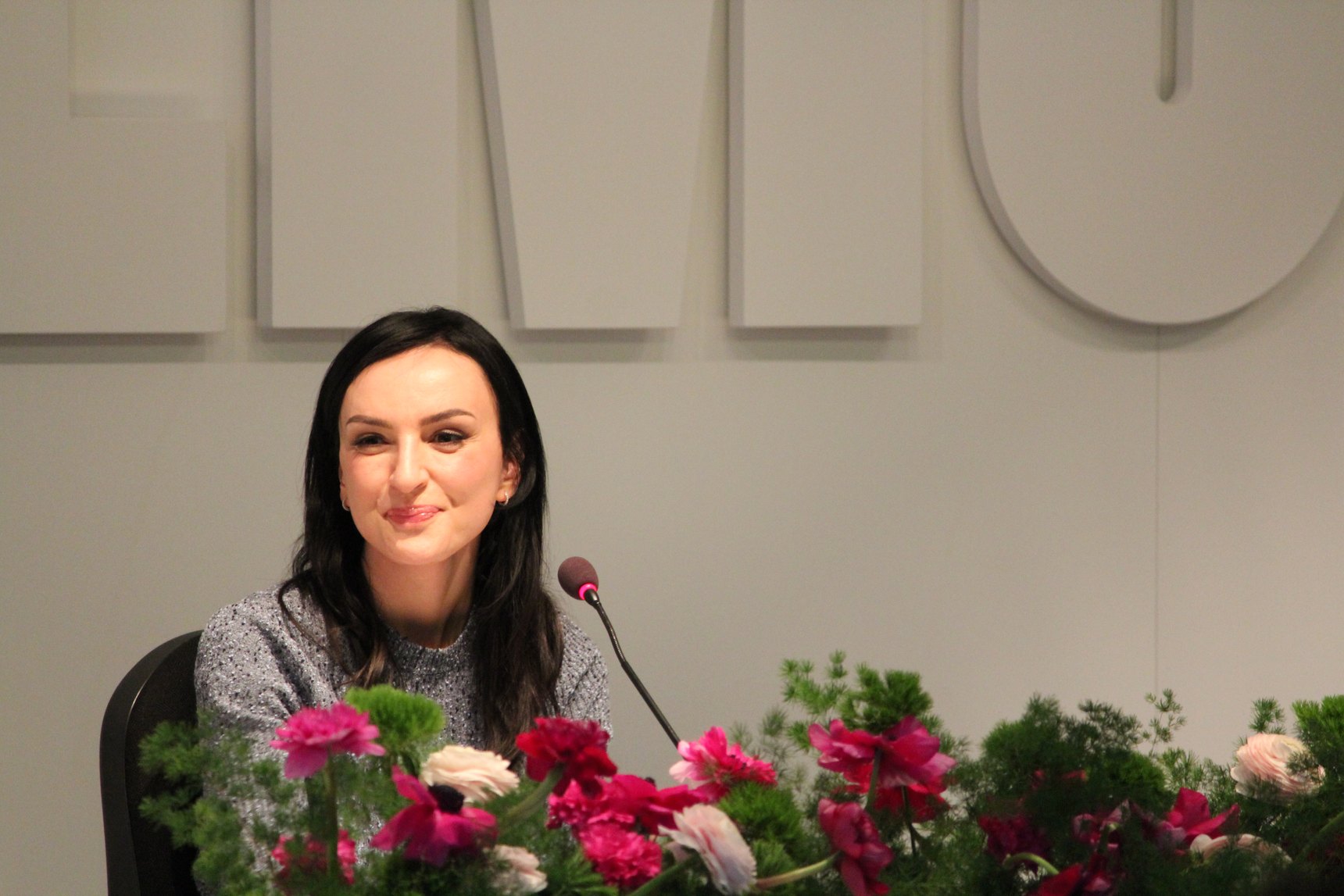 Arisa sorride seduta al tavolo della conferenza stampa di Sanremo, davanti a un microfono e a fiori rosa