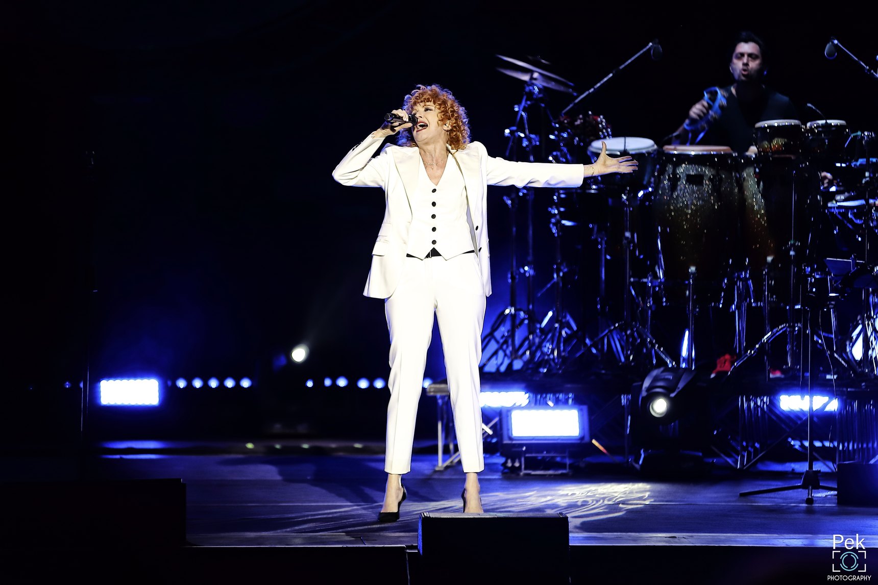 Fiorella Mannoia in completo bianco canta sul palco col braccio teso, alle sue spalle la batteria e luci blu
