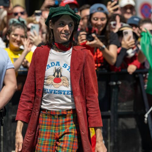 Lucio Corsi in giacca rossa, pantaloni a quadri e cappello verde cammina fra il pubblico, dietro una bandiera italiana