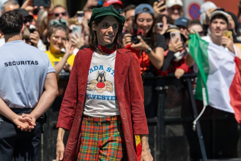 Lucio Corsi in giacca rossa, pantaloni a quadri e cappello verde cammina fra il pubblico, dietro una bandiera italiana