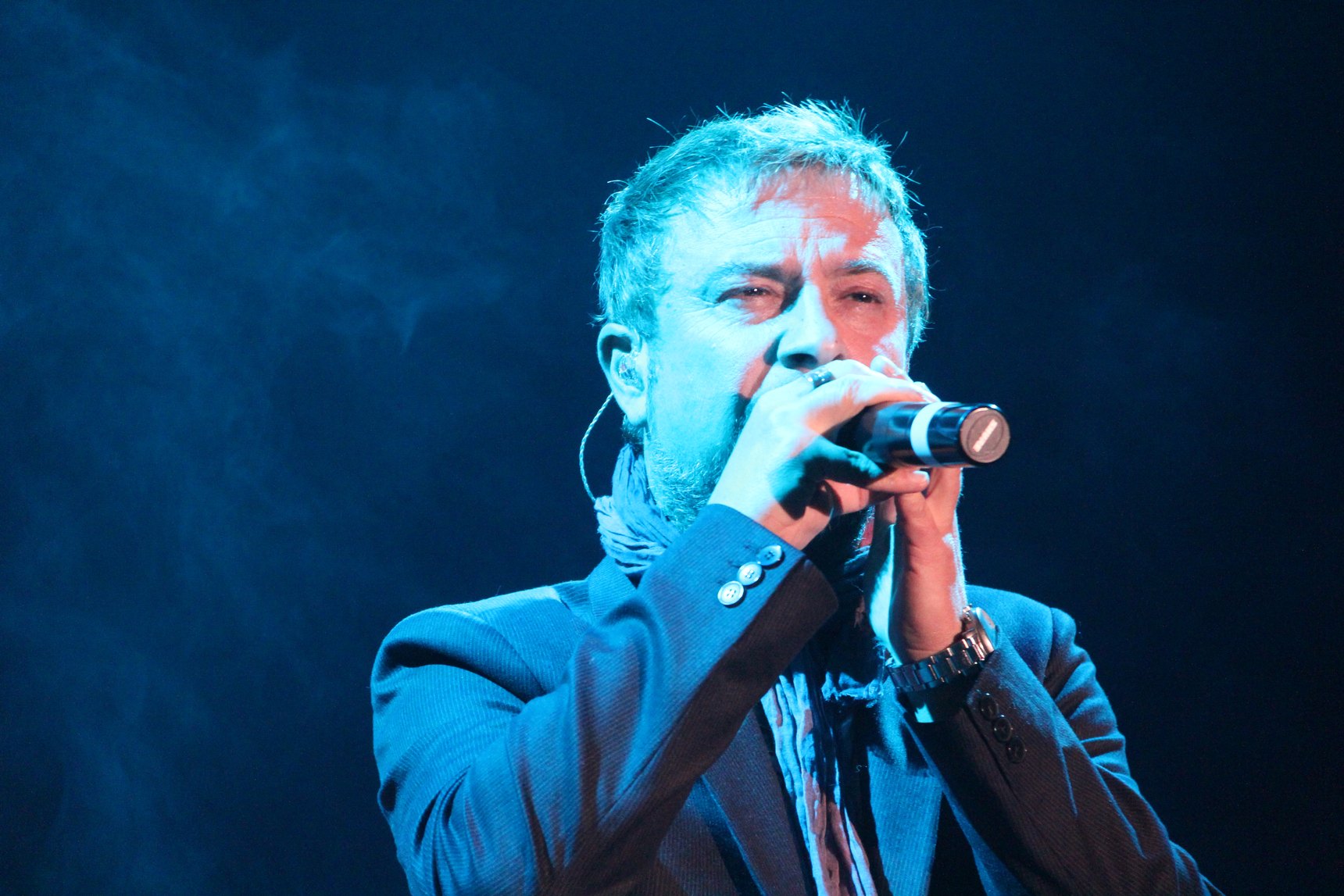 Marco Masini in giacca blu canta stringendo il microfono, illuminato da una luce blu su fondo scuro