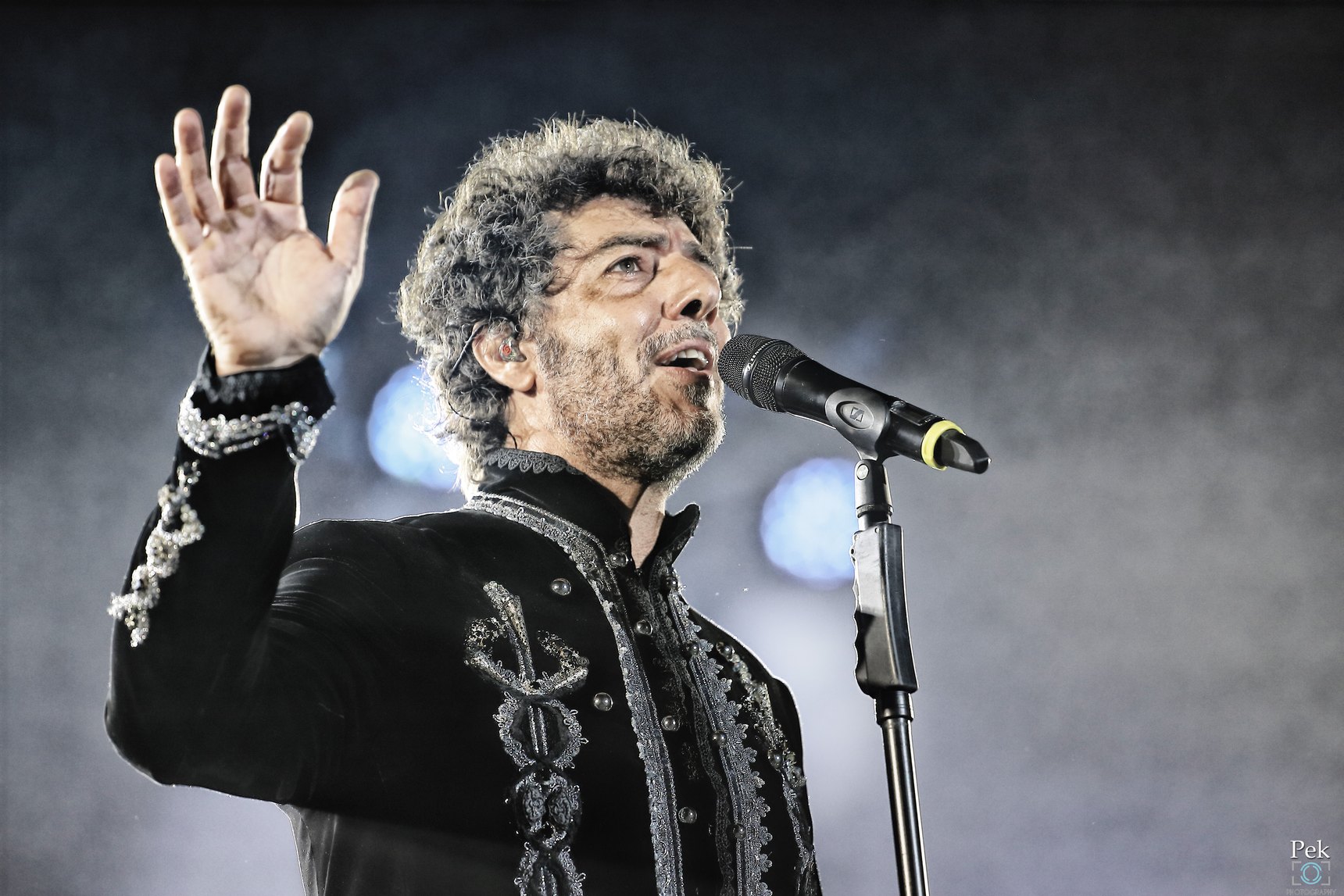 Max Gazzè in giacca nera ricamata canta al microfono con la mano alzata, illuminato su fondo scuro