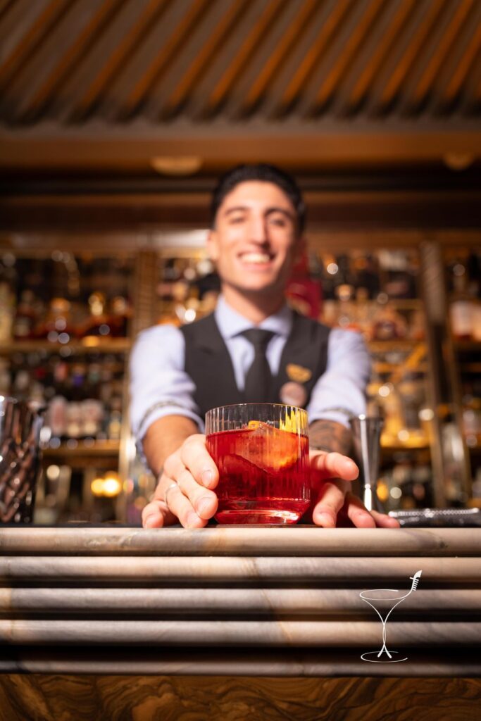 Davide Nasti, sorridente in gilet e cravatta, porge dal bancone un cocktail rosso con la scorza d'arancia