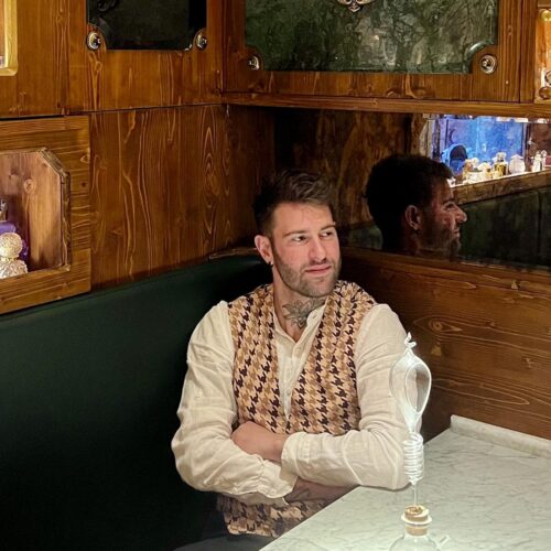 Davide Norcini in camicia bianca e gilet, seduto su una panca verde tra vetrine di flaconi da profumo illuminate