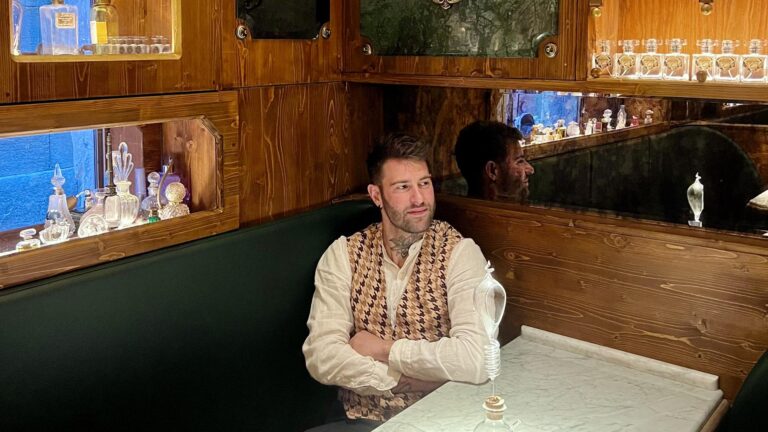 Davide Norcini in camicia bianca e gilet, seduto su una panca verde tra vetrine di flaconi da profumo illuminate