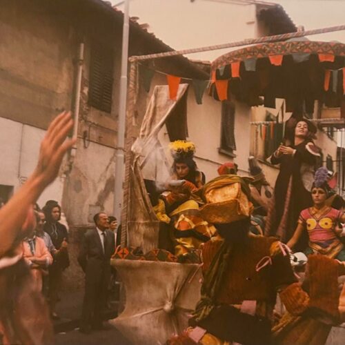 Foto storica a colori di un carro addobbato con festoni e figure in costume che sfila tra le case, con il pubblico ai lati