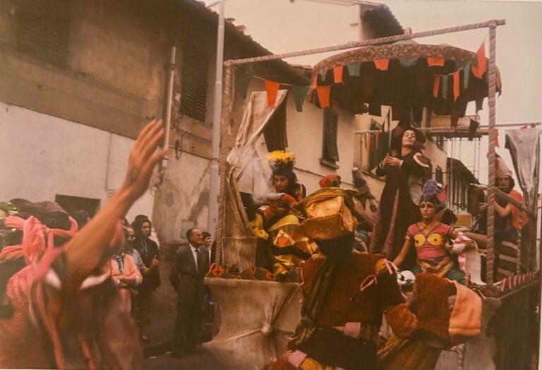 Foto storica a colori di un carro addobbato con festoni e figure in costume che sfila tra le case, con il pubblico ai lati