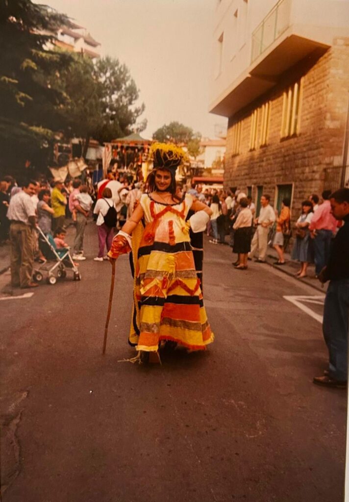 Foto storica di una figura in costume giallo e arancio, con un bastone, che cammina in mezzo alla strada tra la folla