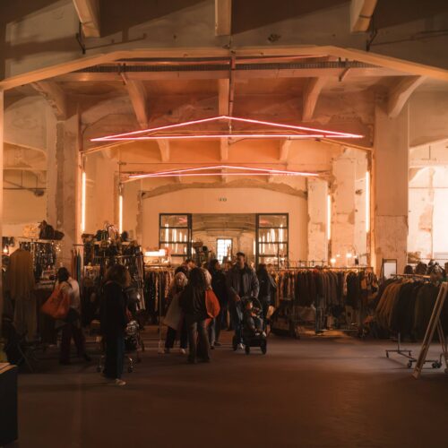 Il padiglione di Manifattura Tabacchi durante East Market: stand di abiti vintage, visitatori e un neon rosa sul soffitto