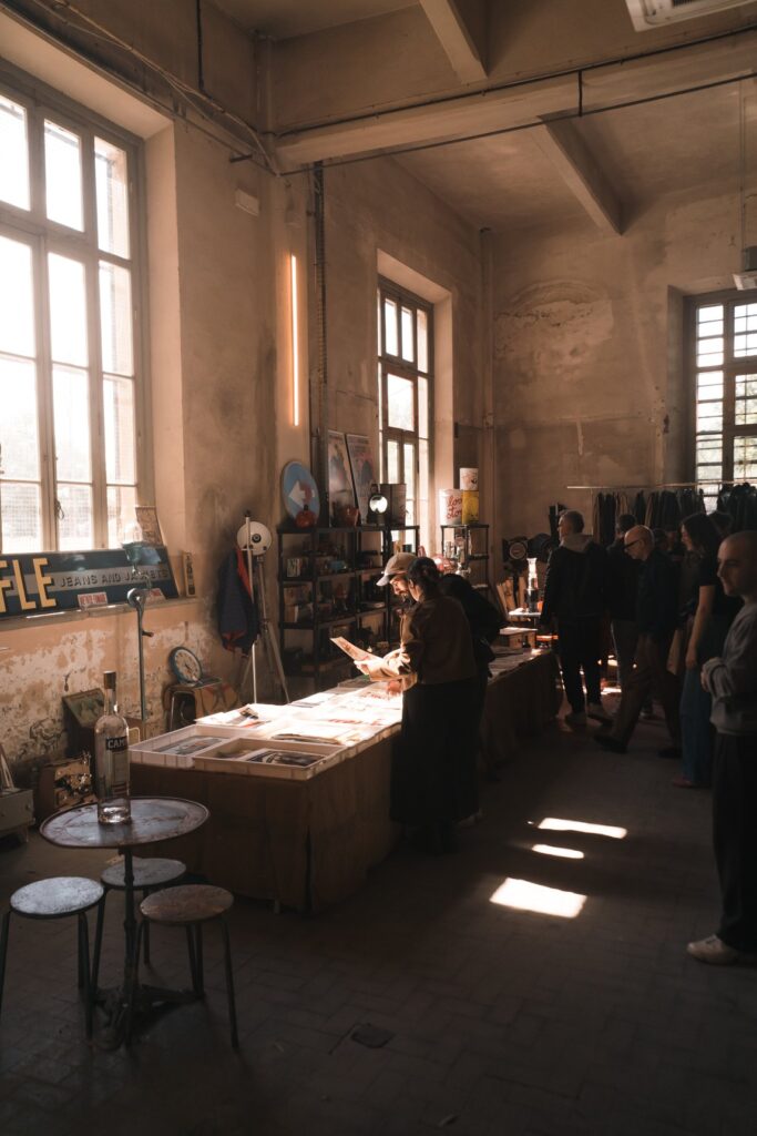 Visitatori che sfogliano stampe e oggetti su un bancone in una sala con grandi finestre di Manifattura Tabacchi