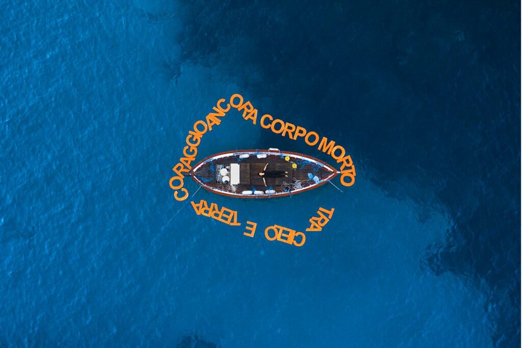 CORPOMORTO di Elena Bellantoni: una barca in mare aperto vista dall'alto, circondata da lettere arancioni galleggianti