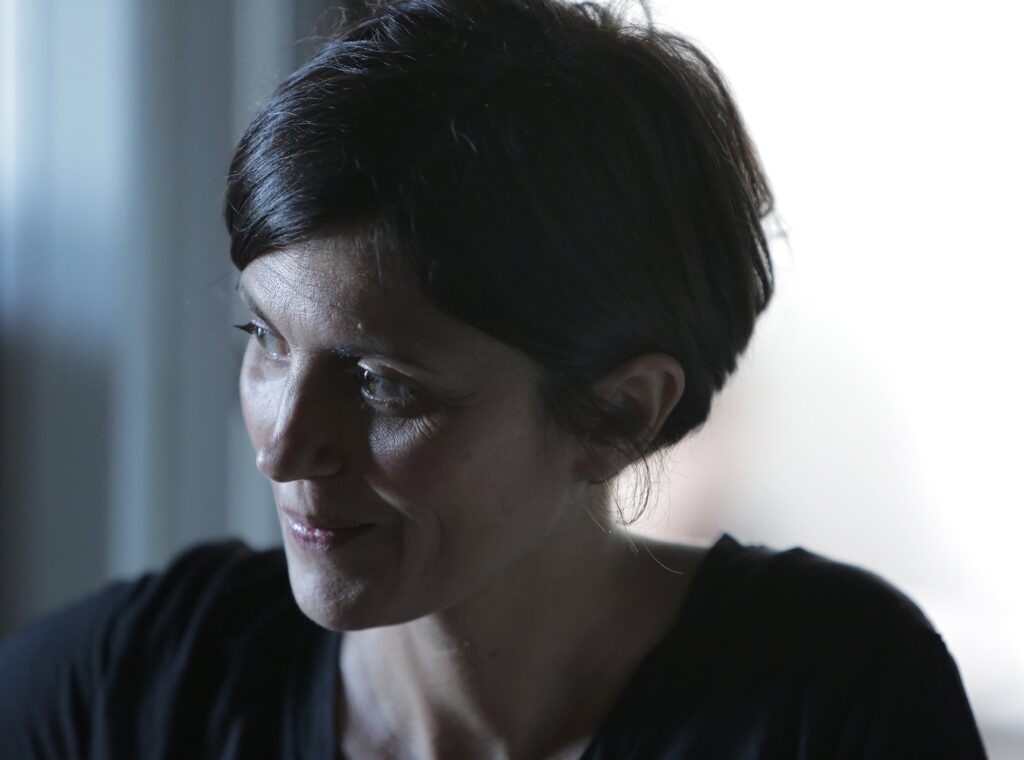 Ritratto dell'artista Elena Bellantoni, capelli corti scuri e maglia nera, sorridente su un fondo chiaro