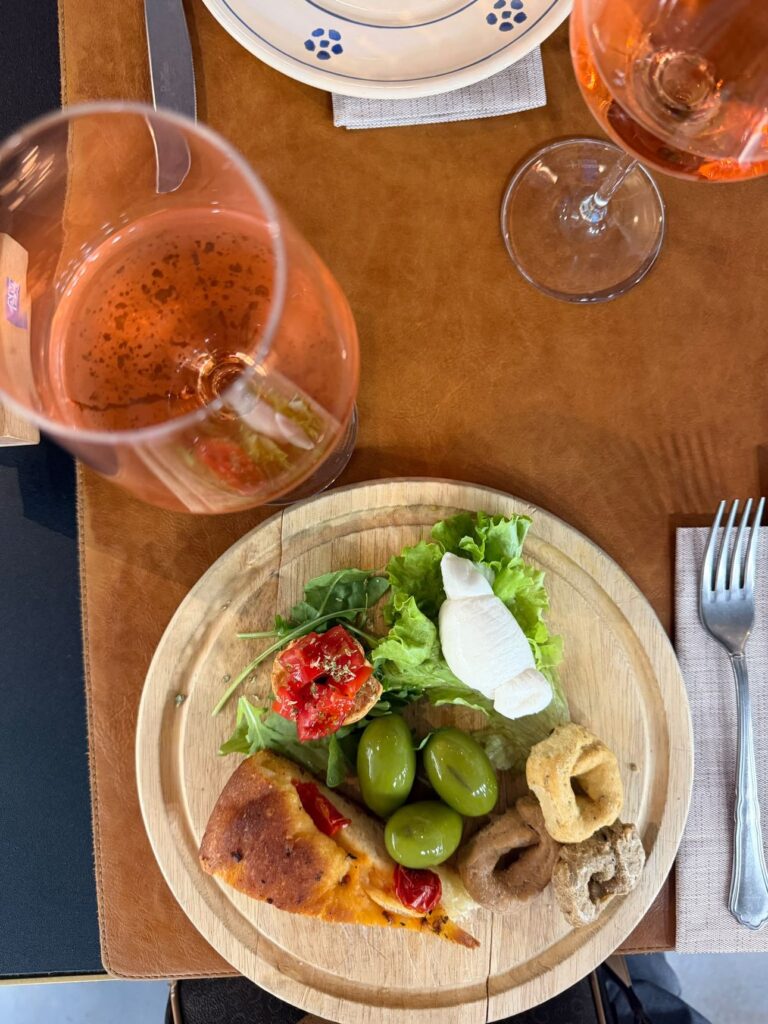 Un tagliere con mozzarella, olive verdi, focaccia e insalata accanto a una caraffa di vino rosato all’Ex Latteria
