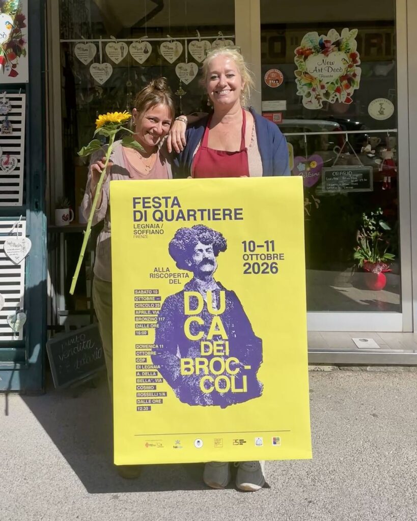 Due donne davanti a un negozio reggono il manifesto giallo e viola della Festa di Quartiere di Legnaia e Soffiano