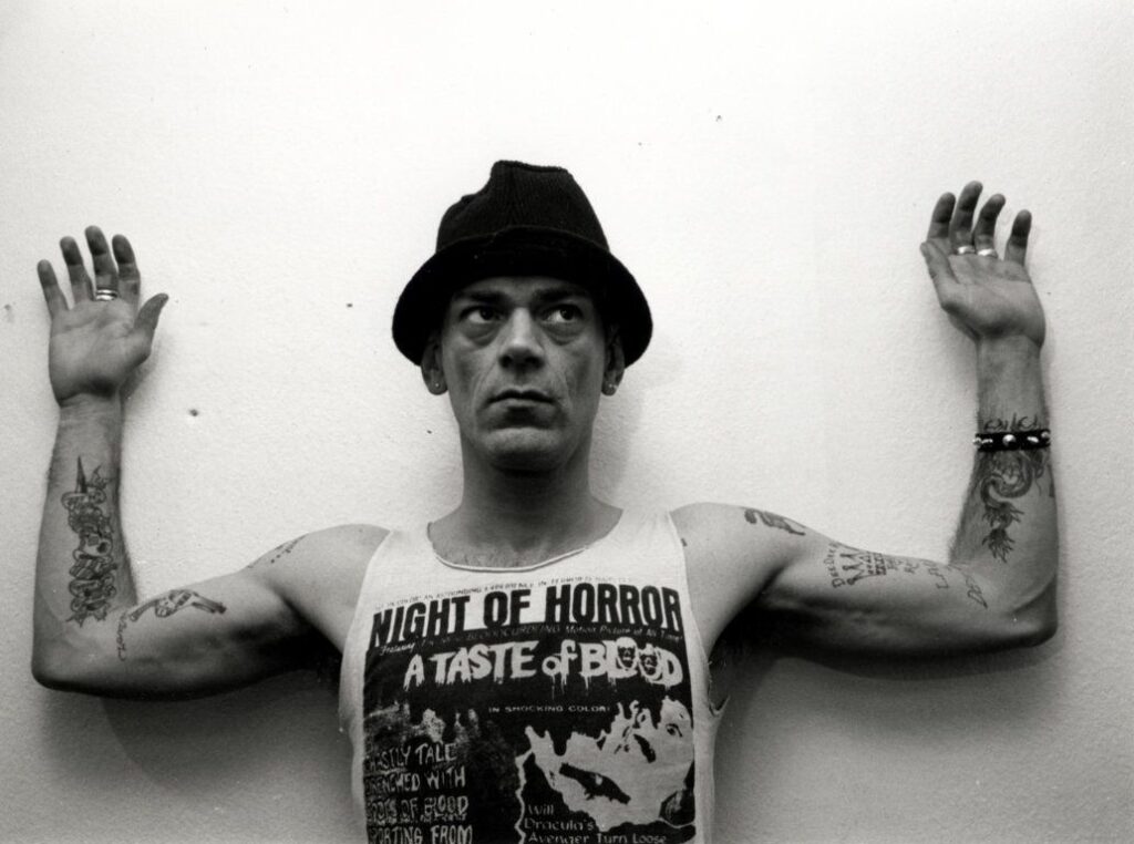 Un ragazzo punk col cappello e le braccia tatuate alzate, con una canottiera «Night of Horror», in bianco e nero