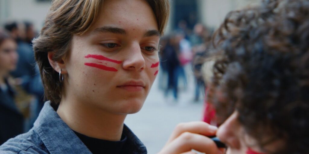 Un ragazzo con due strisce di colore rosso dipinte sulla guancia, di profilo in mezzo a una folla per strada