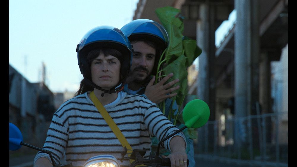 Una donna con la maglia a righe e un uomo dietro di lei, tutti e due col casco blu, su uno scooter in città