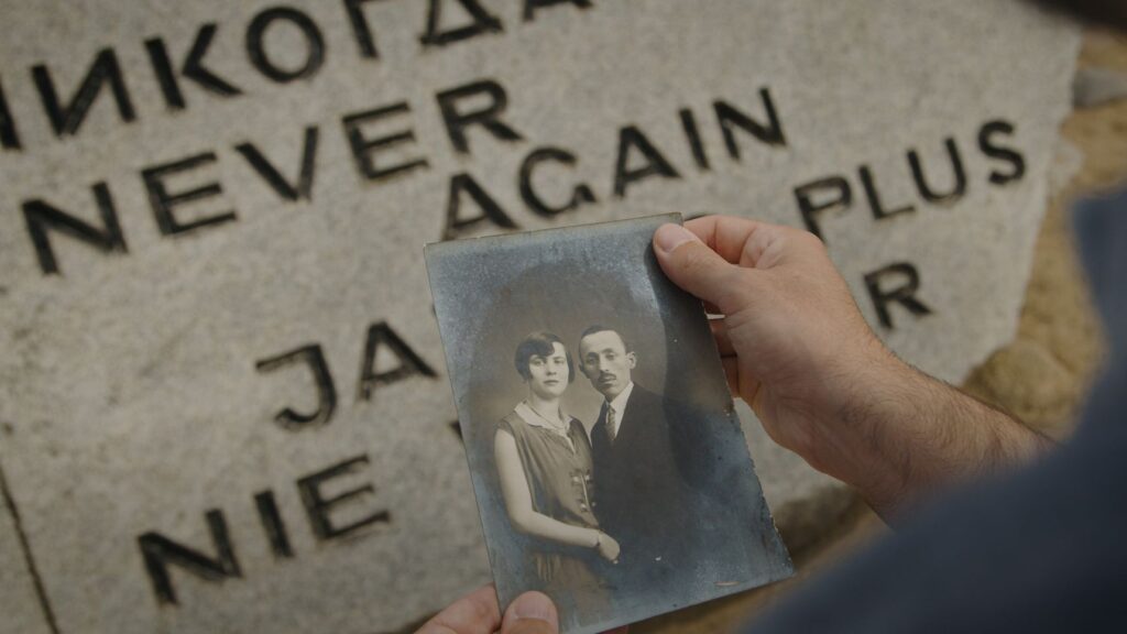 Una mano tiene una vecchia foto di una coppia davanti a una lapide incisa con le parole «Never again»