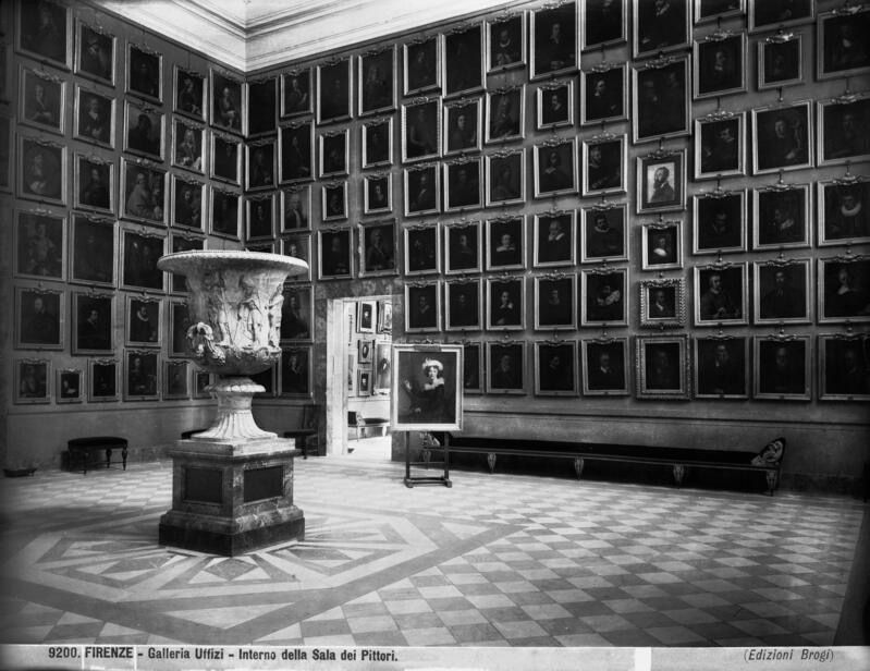 Sala dei Pittori degli Uffizi in una foto d'epoca: pareti coperte di ritratti in cornice, vaso di marmo e pavimento a scacchi