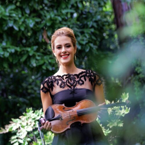 La violinista Francesca Dego in abito nero di pizzo, con il violino in mano, in piedi fra il verde di un giardino