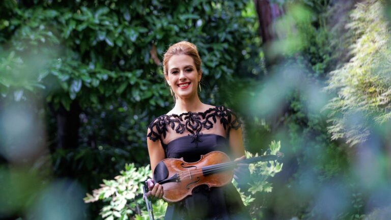 La violinista Francesca Dego in abito nero di pizzo, con il violino in mano, in piedi fra il verde di un giardino