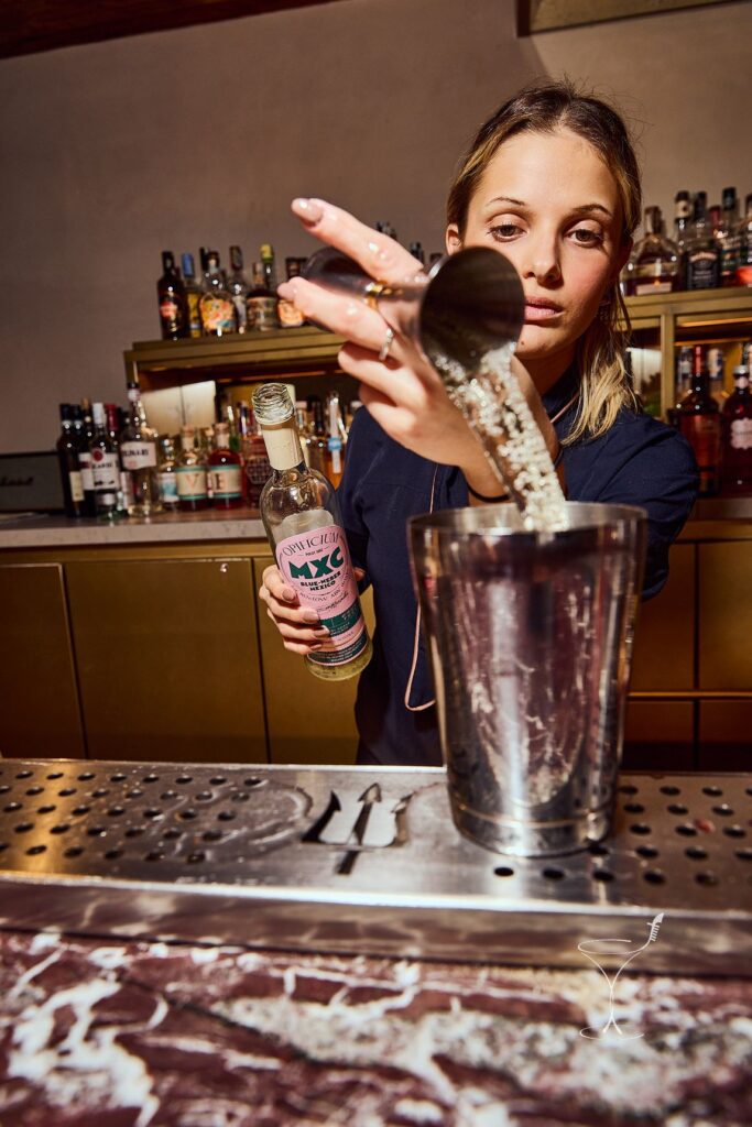 Greta Bazzichi versa dal jigger nello shaker dietro il bancone, con una bottiglia in mano e le bottigliere alle spalle