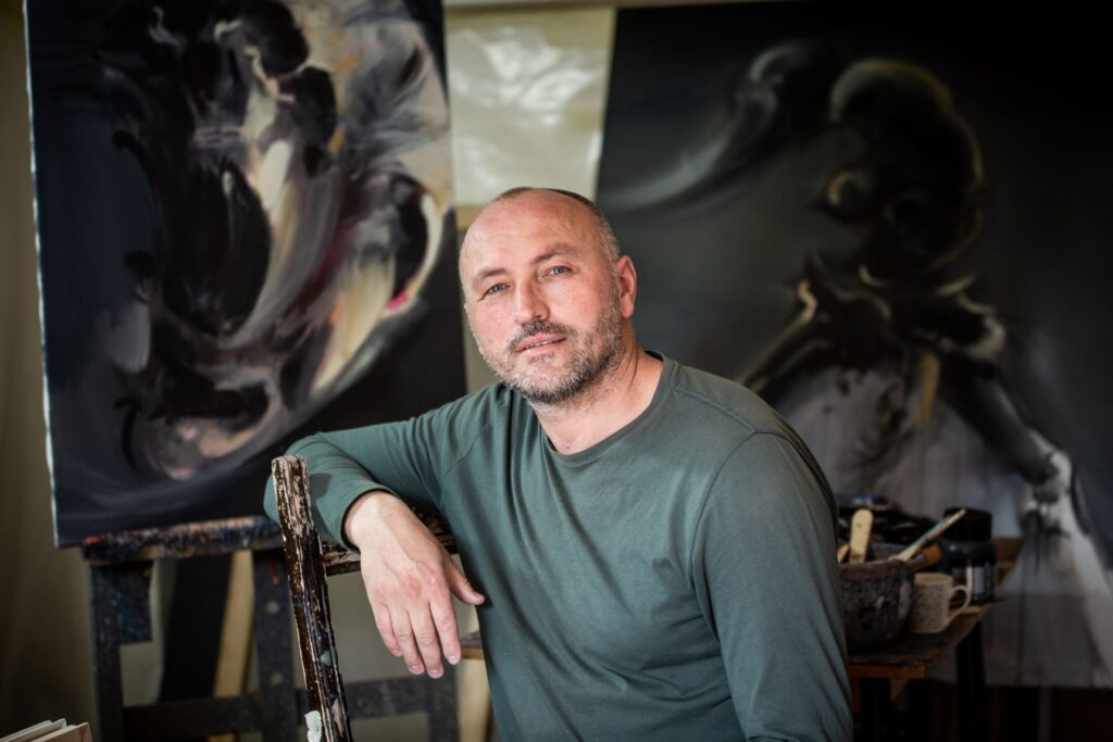 Goran Ćetković, barba corta e maglione verde, seduto in uno studio di pittura davanti a una grande tela scura
