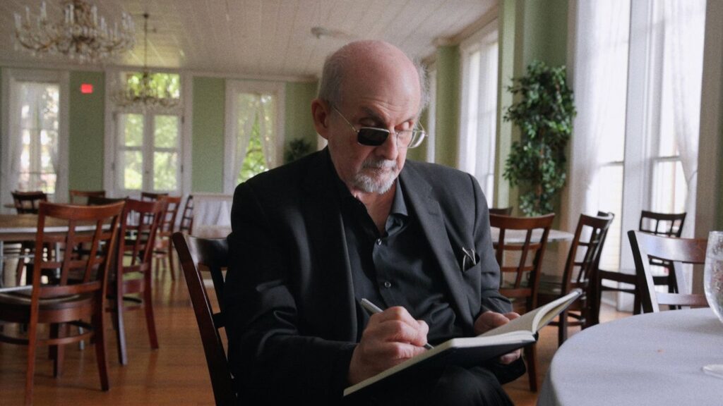 Salman Rushdie, con gli occhiali scuri, scrive su un taccuino seduto al tavolo di una sala con le finestre sul verde