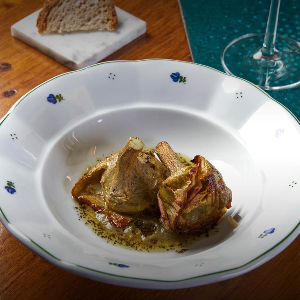 Carciofi alla brace con olio alle erbe in un piatto fondo bianco decorato, accanto a pane e a un calice