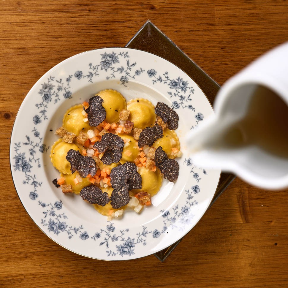 Ravioli con tartufo nero e dadolata di verdure in un piatto a fiori blu, mentre una brocca versa il brodo