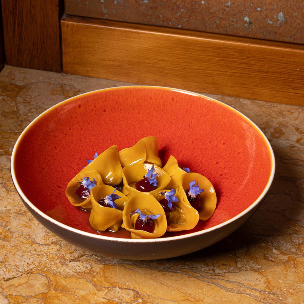 Tortelli gialli con salsa rossa e fiori di borragine blu in una ciotola arancione appoggiata su un piano di marmo
