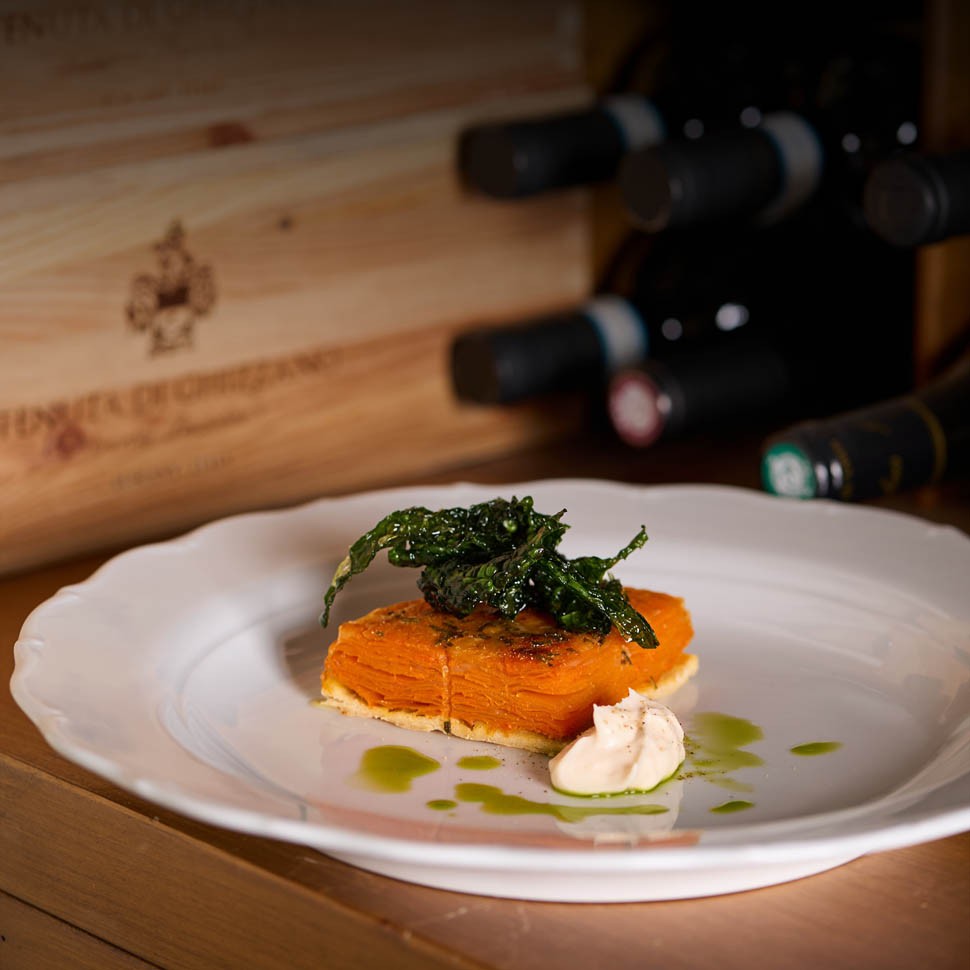 Tartelletta di zucca con cavolo nero croccante e una quenelle chiara su un piatto bianco, davanti a bottiglie di vino