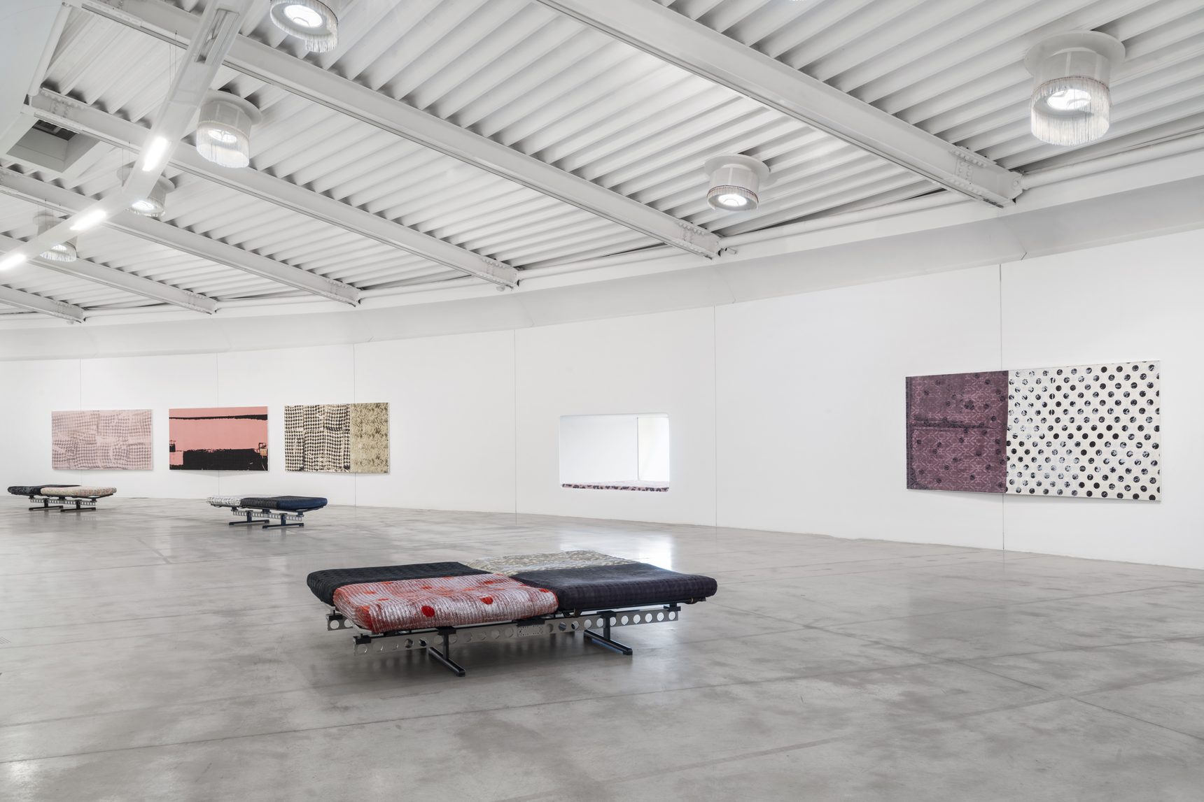 Ampia sala bianca con opere tessili di Lorenza Longhi appese alle pareti e panche rivestite di tessuti stampati