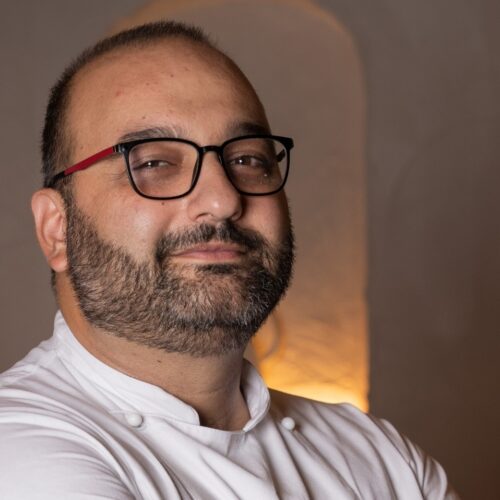 Lorenzo Barsotti in giacca bianca da chef e occhiali, sorride davanti alle nicchie illuminate della sala