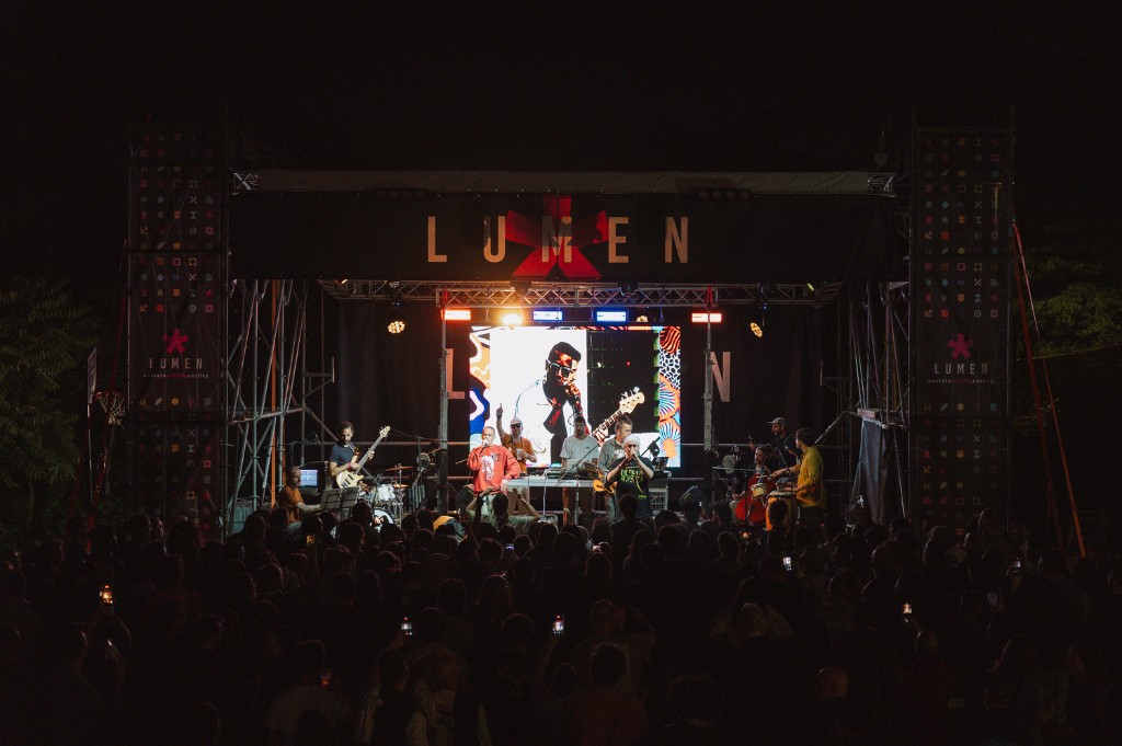 Un concerto di notte sul palco di Lumen, con l'insegna illuminata in alto e il pubblico al buio davanti