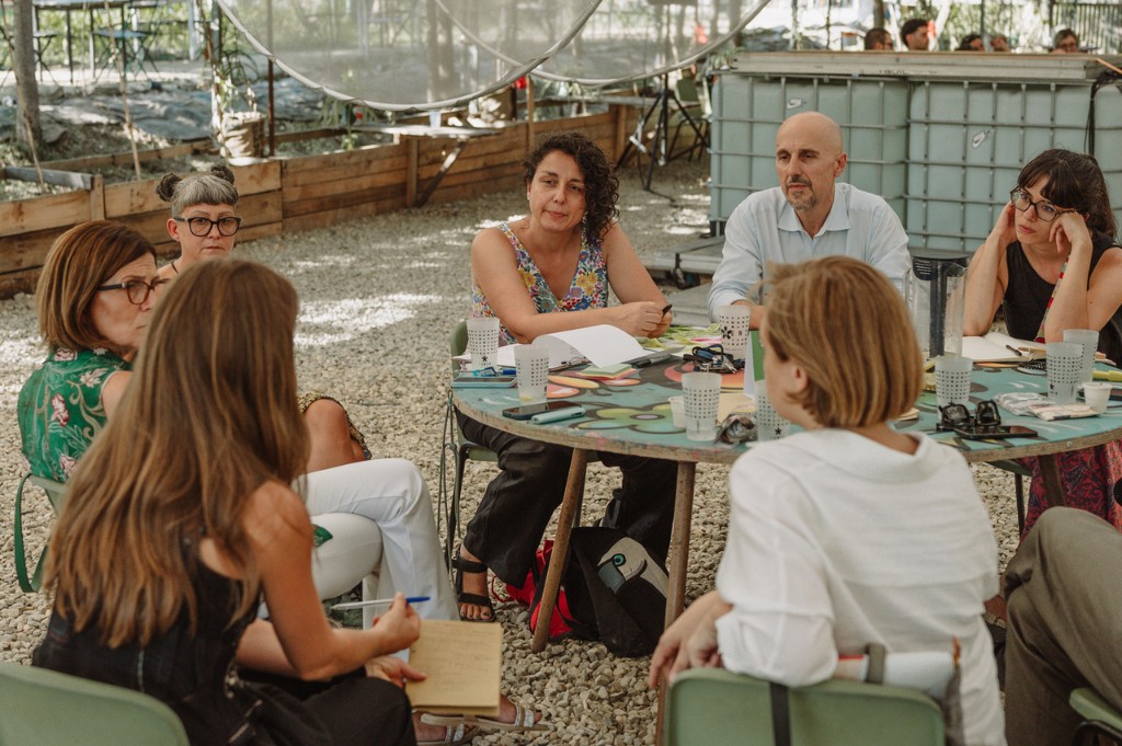 Un gruppo di persone seduto attorno a un tavolo rotondo all'aperto, nel verde di Lumen, durante una discussione