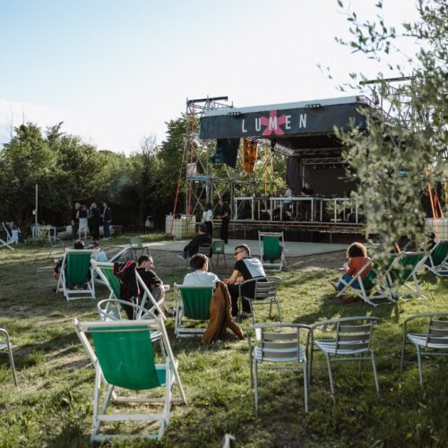 Il palco di Lumen con la grande insegna, di giorno, e davanti il prato con sdraio e sedie tra gli alberi