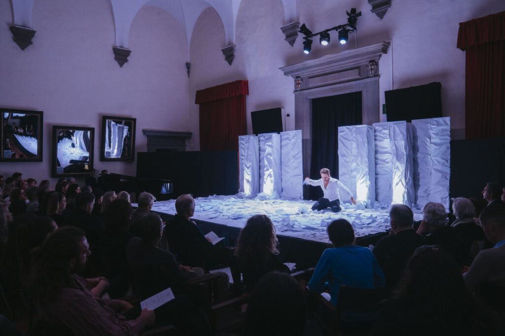 Una cantante seduta su una scena bianca illuminata, davanti a pannelli chiari, con il pubblico in penombra in primo piano